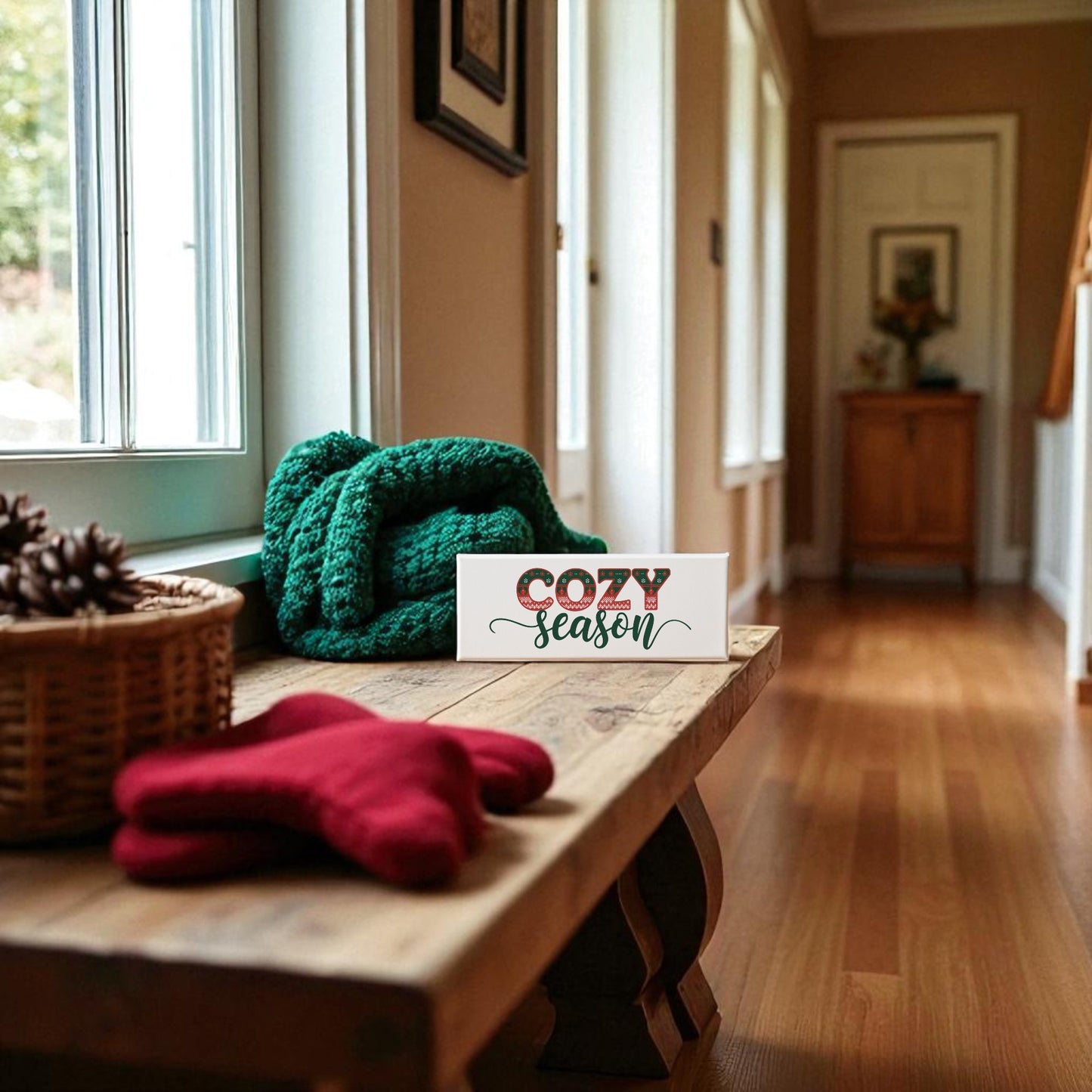 A hallway with wooden floors features a bench holding a green knit blanket, red mittens, pinecones in a basket, and the Cozy Season Canvas Sign (4x12) by Mirabilia Boutique. Sunlight streams through large windows, creating an inviting holiday scene.