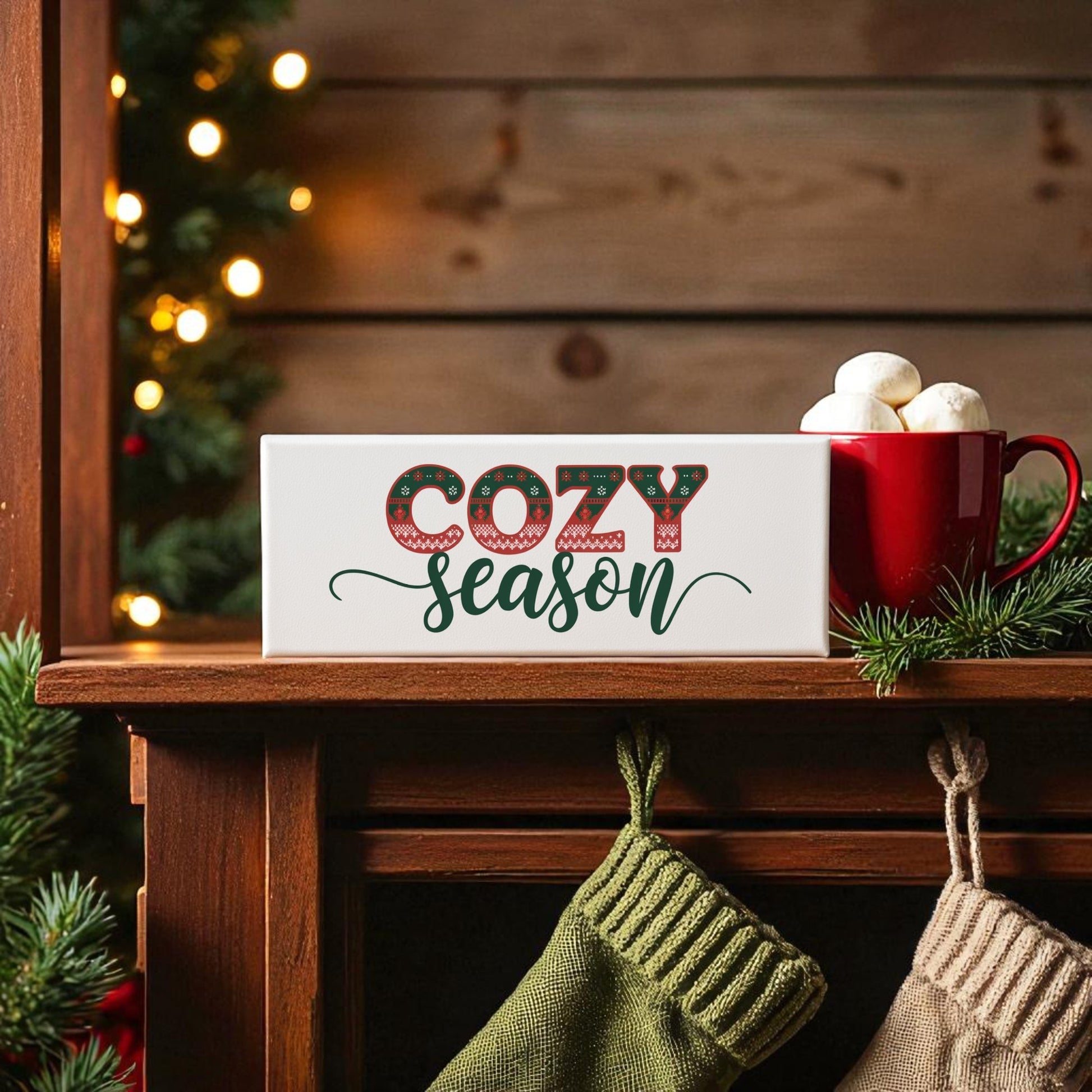 A festive mantel with stockings, a red mug with marshmallows, pine branches, and the Mirabilia Boutique Cozy Season Canvas Sign (4x12 holiday wall decor) glows warmly—perfect holiday home decor against a wooden backdrop.