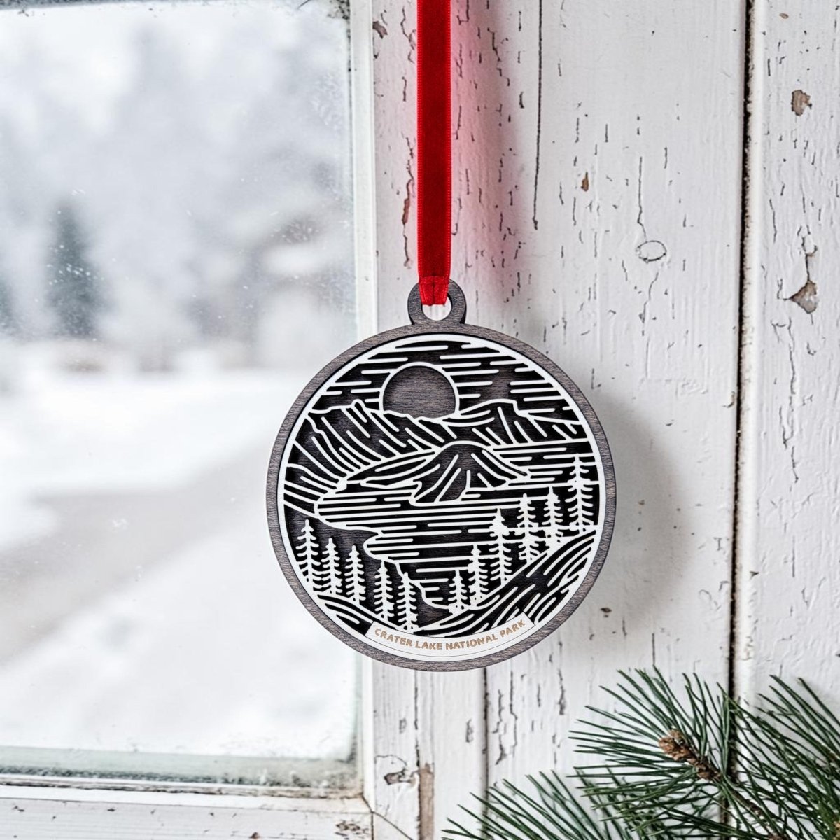 The Crater Lake National Park Christmas Ornament by Mirabilia Boutique, featuring oval wooden line art of a mountain and forest scene with "Crater Lake National Park" inscribed, hangs from a red ribbon on a white window. Snowy trees are visible outside, while pine branches appear in the lower right foreground.