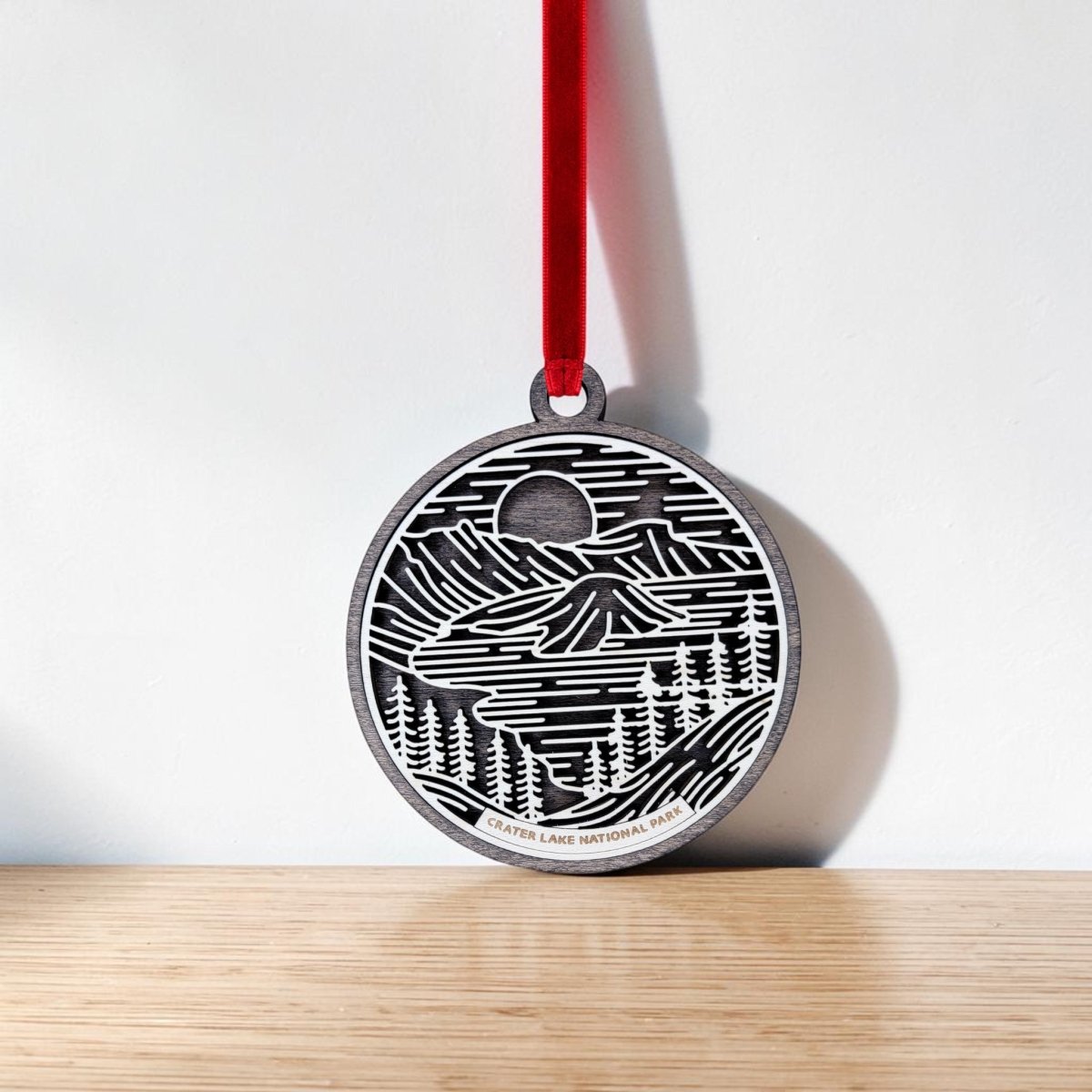 A Crater Lake National Park Christmas Ornament by Mirabilia Boutique, featuring a round wooden design with an engraved black and white landscape, hangs from a red ribbon against a white wall above a wooden surface, highlighting the park’s beauty in elegant line art style.