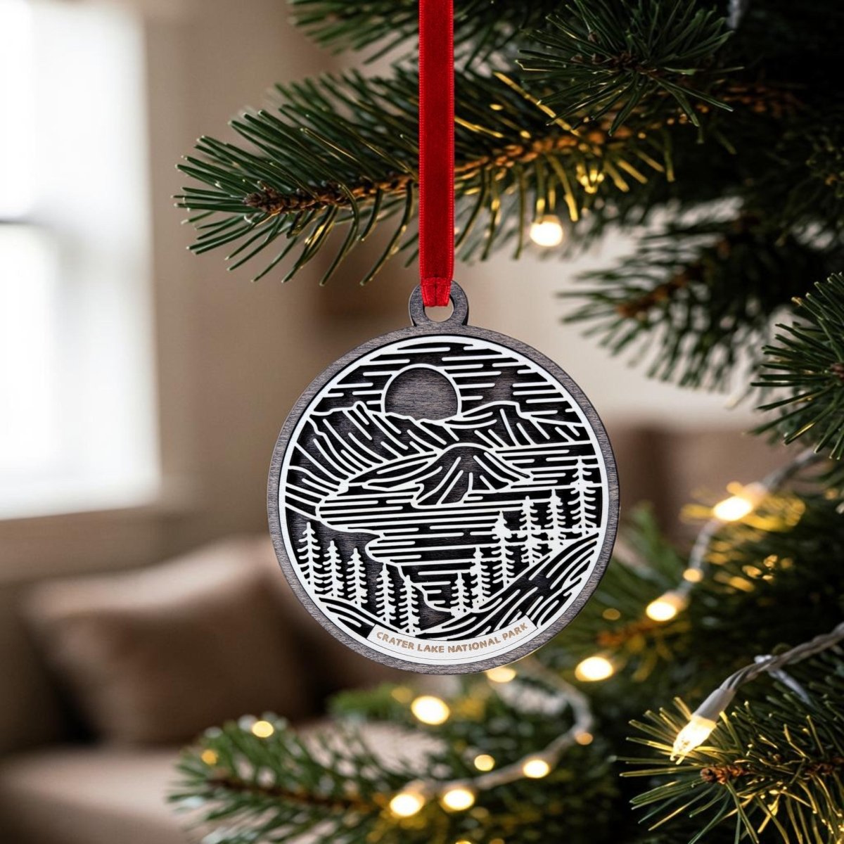 A Crater Lake National Park Christmas Ornament by Mirabilia Boutique, featuring a mountain and trees design in wooden line art, hangs from a red ribbon on a Christmas tree decorated with lights. With a blurred sofa in the background, this beautiful ornament captures the serene landscape of Crater Lake, making it a unique holiday keepsake.