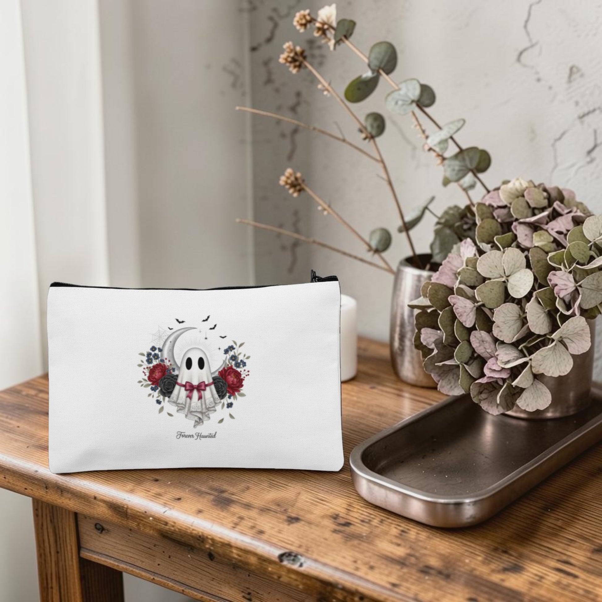 The Forever Haunted Makeup Bag | Gothic Ghost Pouch by Mirabilia Boutique, featuring a ghost and floral design in white, is displayed on a wooden table beside a dried hydrangea arrangement in a metallic tray and a minimalist vase with branches, all set against a textured wall.