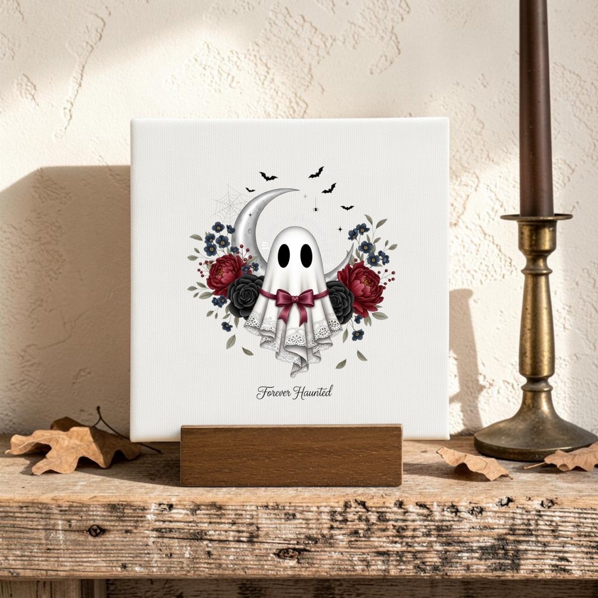 The Forever Haunted Mini Canvas Sign by Mirabilia Boutique features a charming ghost with a red bow, lace accents, and delicate flowers, set against a backdrop of crescent moon and bats. The phrase "Forever Haunted" is displayed below the design. Perfect for Halloween home decor or adding gothic cottagecore vibes to your space, this mini canvas sits on a wooden stand beside a candle and autumn leaves.