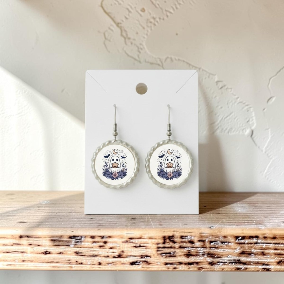 A pair of Crescent Moon Pumpkin Ghost Scalloped Earrings by Mirabilia Boutique, featuring whimsical pumpkin and ghost designs with scalloped edges, hang on a white card, displayed on a wooden shelf against a textured white wall.