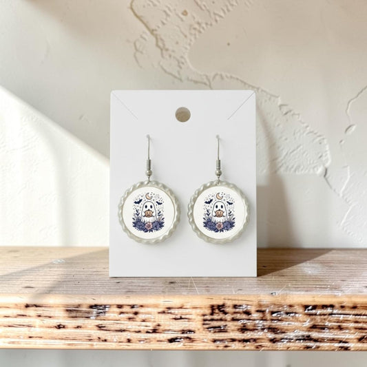 A pair of Crescent Moon Pumpkin Ghost Scalloped Earrings by Mirabilia Boutique, featuring whimsical pumpkin and ghost designs with scalloped edges, hang on a white card, displayed on a wooden shelf against a textured white wall.