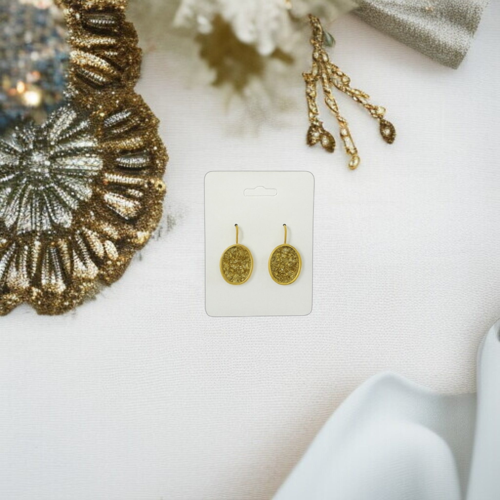 Mirabilia Boutique’s Gold Crushed Glass Earrings | Sparkling Oval Dangle Jewelry are shown on a white card placed on white fabric with gold accents and a blue cloth in the corners. Minimalist hook design in elegant gold.