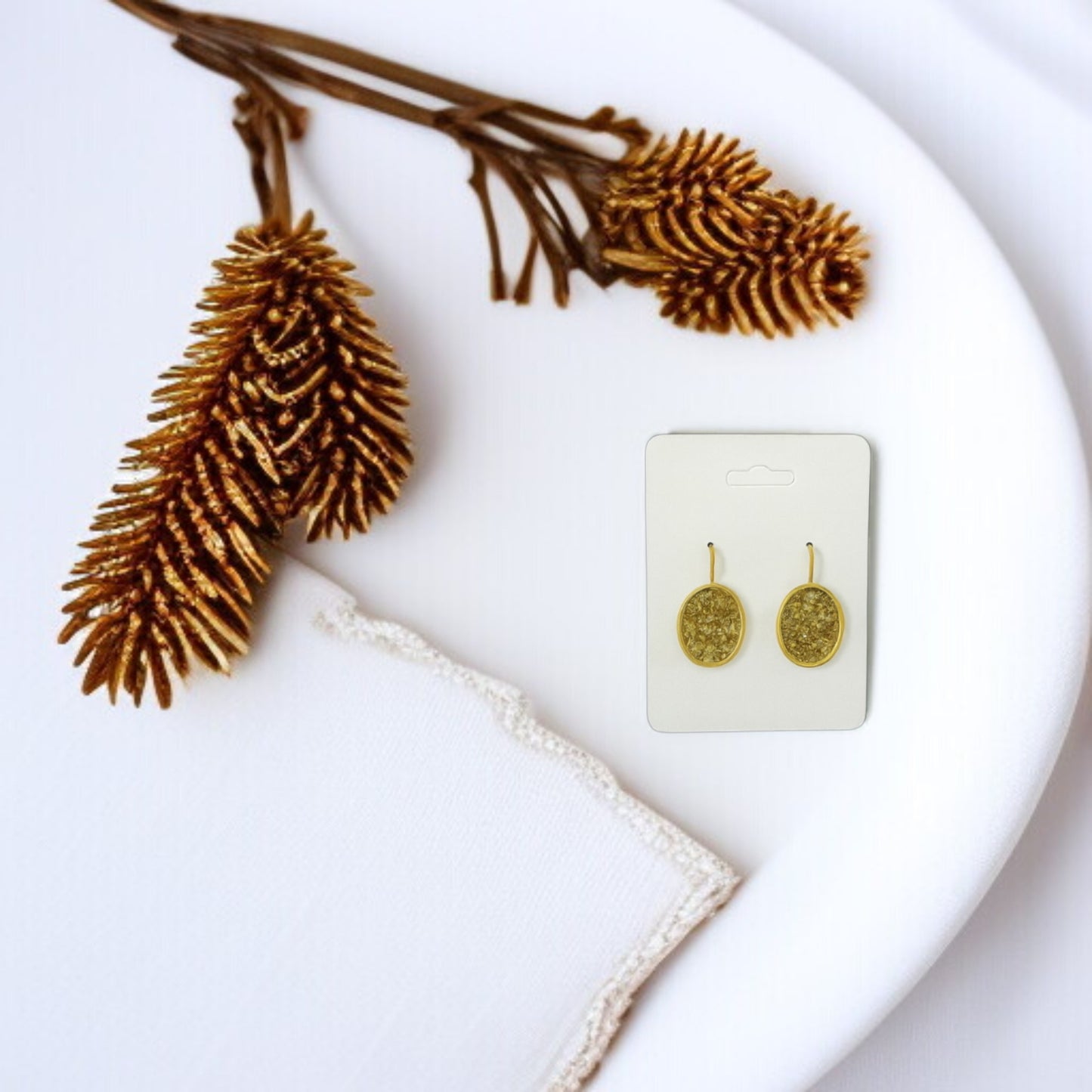 The Gold Crushed Glass Earrings by Mirabilia Boutique—a pair of sparkling oval dangle jewelry—is shown on a display card atop a white plate, next to pinecones and a decorative-edged white cloth.