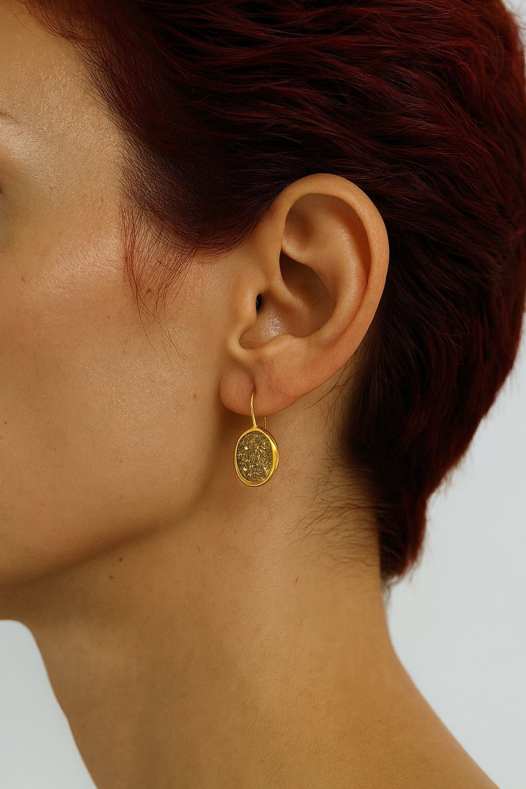 Close-up of a person with short, dark red hair wearing Mirabilia Boutique's Gold Crushed Glass Earrings—minimalist hooks with sparkling oval dangles—set against a plain light background, showing the ear, part of the face, and neck.