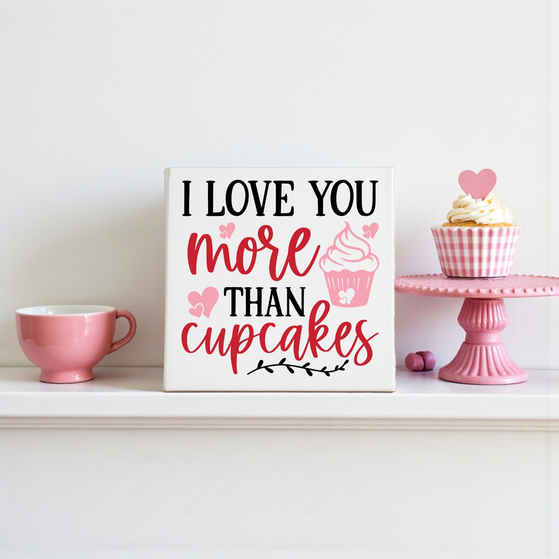 A pink cup, a cupcake with a heart topper on a pink stand, and the Mirabilia Boutique "I Love You More Than Cupcakes" 8x8 Canvas Sign add sweet Valentine’s decor to the white shelf.