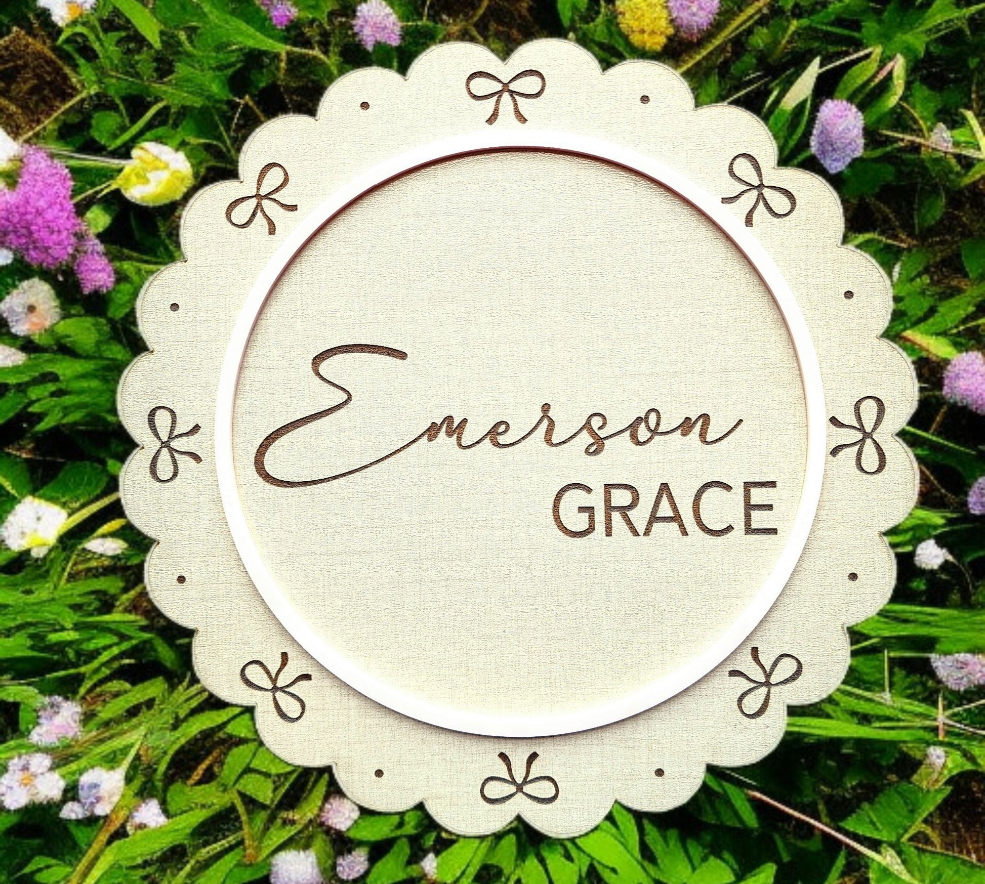 The Personalized Baby Name Shelf Sign by Mirabilia Boutique features a round, scalloped edge with bows and “Emerson Grace” in script—perfect nursery shelf decor set against green leaves and pink, white, and purple flowers.