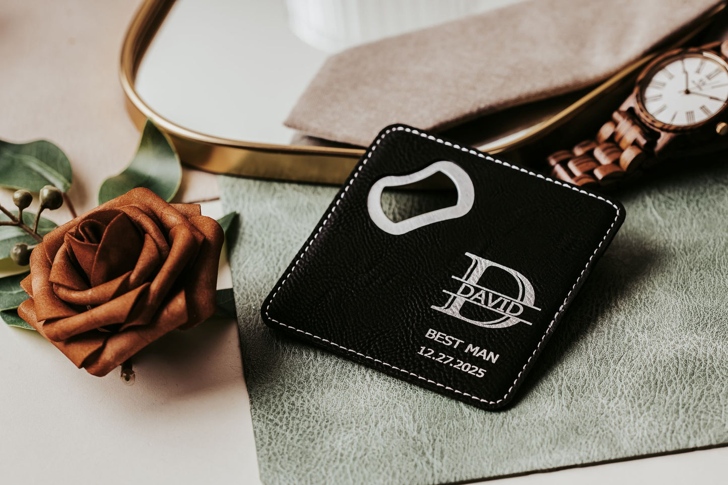 The Engraved Leatherette Bottle Opener Coaster by Mirabilia Boutique, personalized with "David," "Best Man," and "12.27.2025," sits on a green cloth near a wooden watch, brown rose, and gold-edged mirror—a unique groomsman gift.
