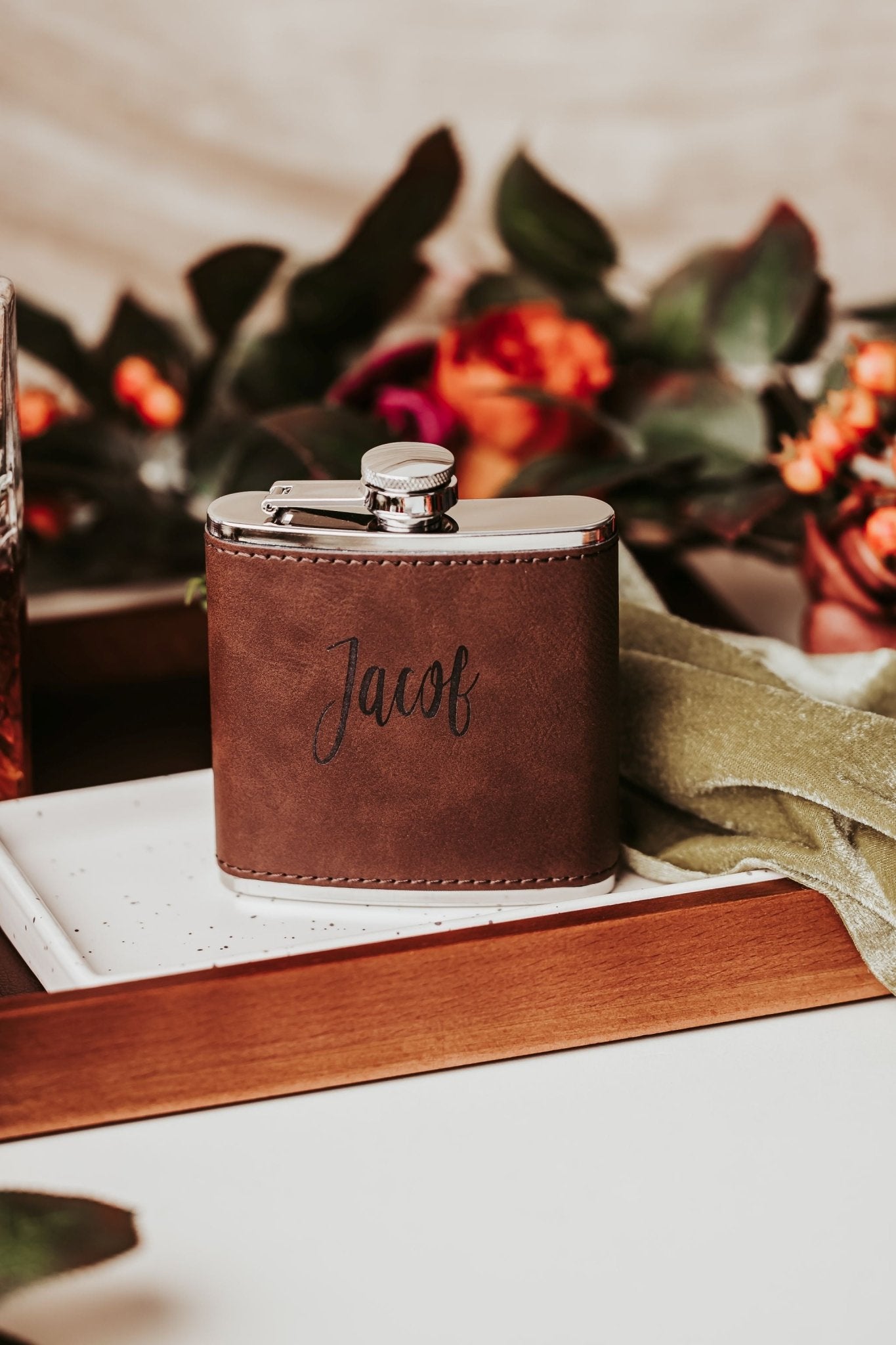 The Custom Engraved Flask by Mirabilia Boutique, wrapped in leather and personalized for him, rests on a tray amid flowers—making it the perfect groomsmen gift for any special occasion.