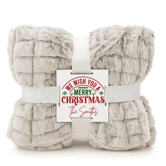 A soft beige faux fur Custom Christmas Throw Blanket by Mirabilia Boutique is folded, tied with a white ribbon, and includes a card reading "We wish you a Merry Christmas" with "The Smiths" in red—perfect cozy holiday gift!.