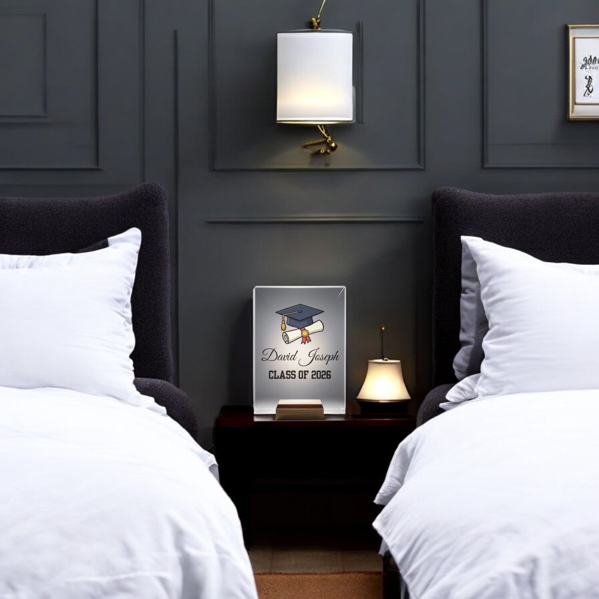 The Classic Graduation Cap Personalized Night Light by Mirabilia Boutique, engraved with "David Joseph CLASS OF 2026" and a graduation cap, sits on a nightstand between two white-bedded, gray-headboard beds in a modern, dark-walled room.