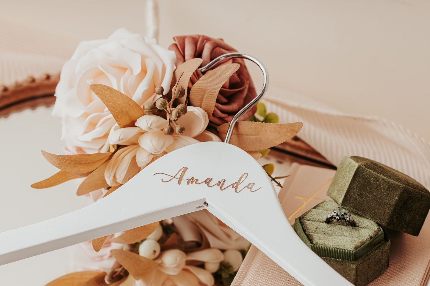 A Mirabilia Boutique Personalized Wedding Hanger from the Custom Name Hanger Set, labeled “Amanda,” is displayed beside blush and beige artificial flowers, a green velvet ring box with a diamond ring, and a mirror in the background.