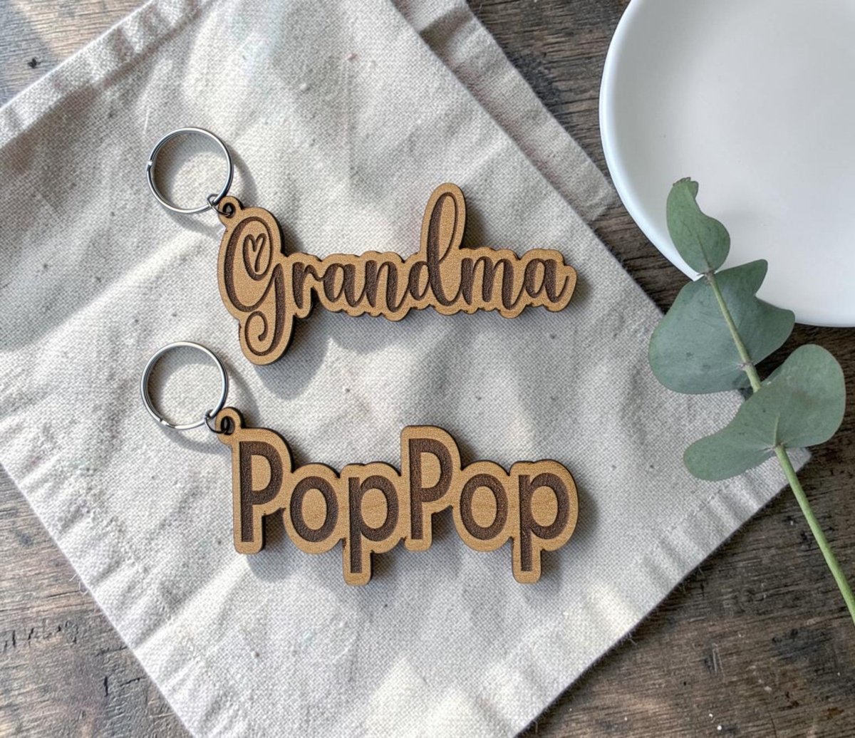 Two Personalized Engraved Wood Name Keychains from Mirabilia Boutique rest on a beige cloth. One keychain is engraved with "Grandma," and the other with "PopPop." A small white plate and green eucalyptus leaves nearby on the wooden surface accentuate the charm of each custom name keychain.