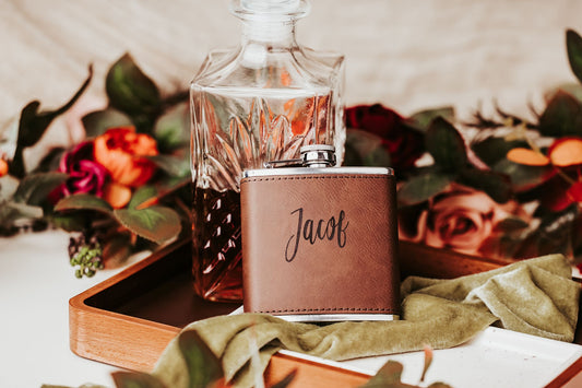 The Custom Engraved Flask by Mirabilia Boutique, a personalized leather gift for him, rests on a wooden tray with a glass decanter, green velvet cloth, and colorful flowers—a perfect groomsmen gift.