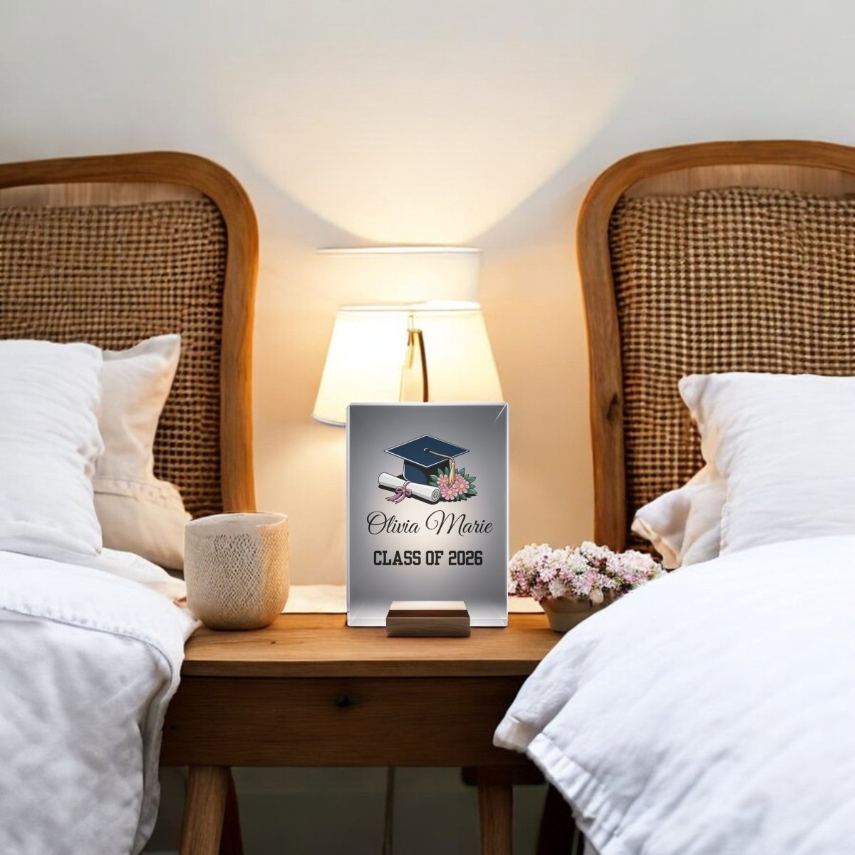 A wooden nightstand between two beds holds a lamp, a white mug, flowers, and the Mirabilia Boutique Floral Personalized Graduation Night Light featuring "Olivia Marie, Class of 2026" with a graduation cap and diploma illustration.