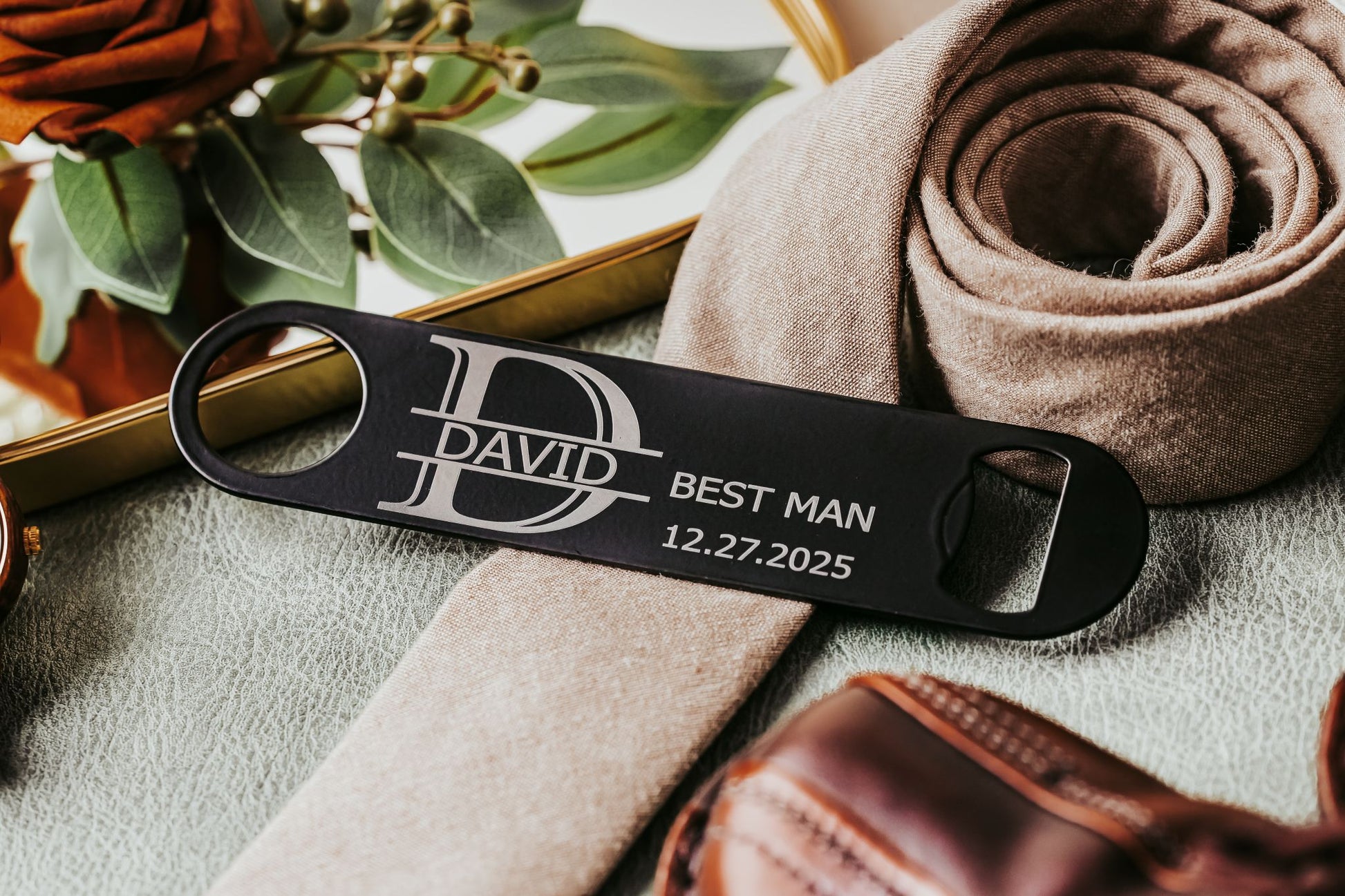 The Mirabilia Boutique Engraved Flat Metal Bottle Opener, etched with "DAVID BEST MAN 12.27.2025," rests among elegant decor—a classy groomsmen gift or custom wedding favor for your big day.
