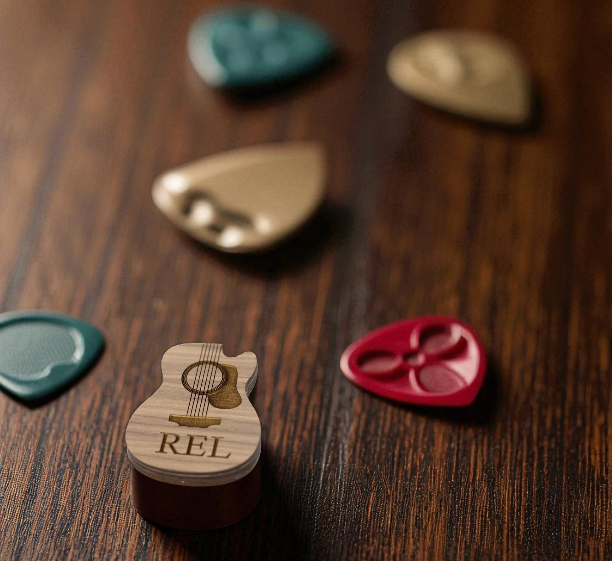 The Custom Guitar Pick Box by Mirabilia Boutique, a small wooden holder shaped like a guitar and engraved with "REL," rests on a wooden surface surrounded by colorful guitar picks—a perfect personalized music gift for guitarists.