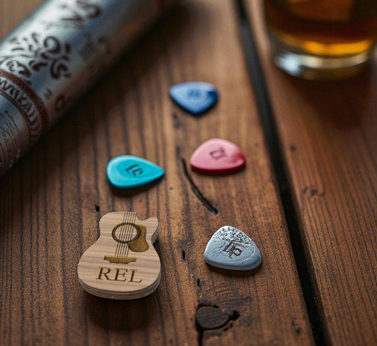 The Custom Guitar Pick Box by Mirabilia Boutique, engraved with "REL" and shaped like a guitar, sits on a wooden table beside four colorful picks, a patterned tube, and a drink in the background—a perfect personalized gift for guitarists.