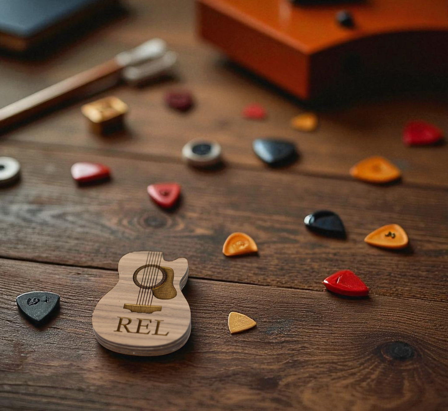 The Mirabilia Boutique Custom Guitar Pick Box, a personalized music gift for guitarists, sits on a wooden table with colorful picks around it and a blurry orange instrument in the background.