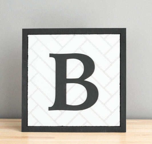 The Mirabilia Boutique Custom Initial Shelf Sign | Herringbone Backer | 10x10 features a bold black "B" on white with a herringbone pattern—ideal for personalized modern farmhouse decor on shelves or mantels.