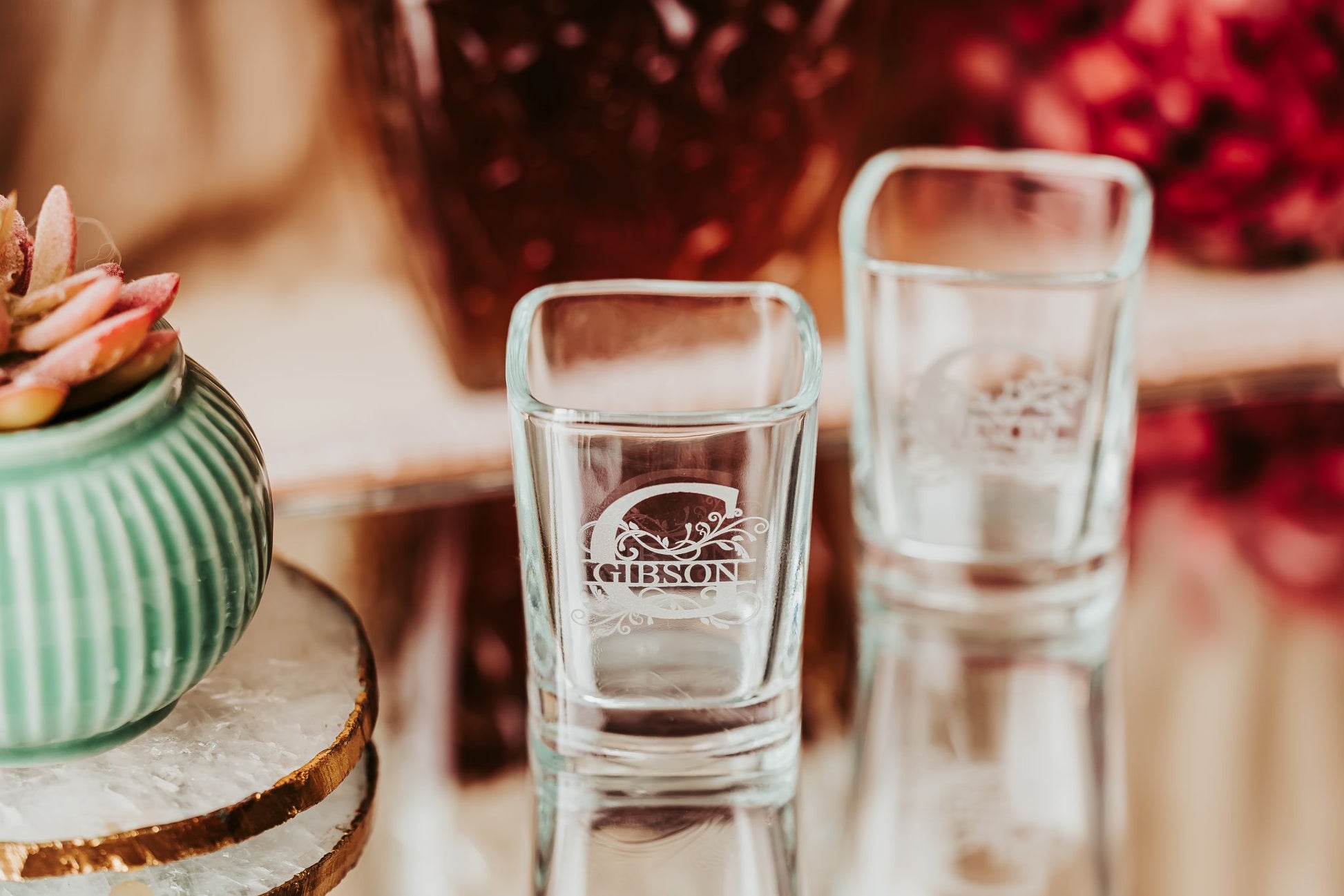 Two Personalized Monogram Shot Glasses by Mirabilia Boutique, custom engraved with "GIBSON" and a decorative design, rest on a reflective surface beside a green ribbed ceramic succulent planter. The softly blurred background adds style.