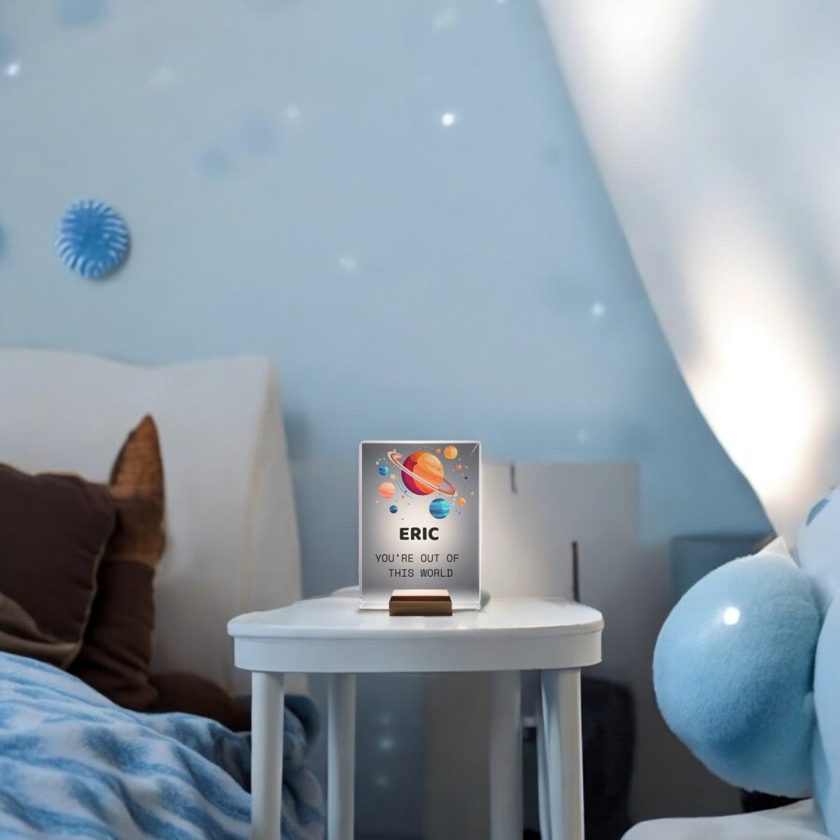 A Mirabilia Boutique Personalized Space Night Light for Kids with colorful planets and a plaque reading “ERIC YOU’RE OUT OF THIS WORLD”—perfect for a cozy, softly lit blue space-themed bedroom with comfy bedding.
