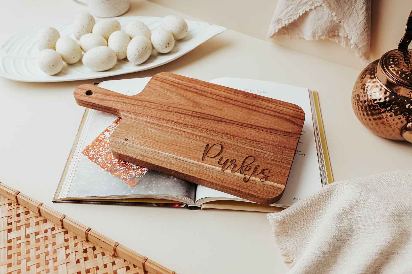 The Mirabilia Boutique Engraved Acacia Cutting Board | 14x8 Personalized Gift, custom-engraved with "Purkis," sits atop an open cookbook, beside speckled eggs, a copper teapot, and neutral textiles on a beige surface.