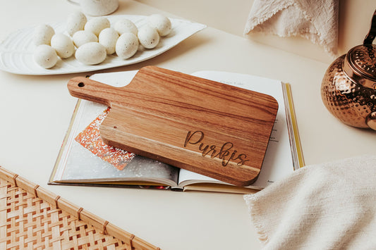 The Mirabilia Boutique Engraved Acacia Cutting Board | 14x8 Personalized Gift, custom-engraved with "Purkis," sits atop an open cookbook, beside speckled eggs, a copper teapot, and neutral textiles on a beige surface.