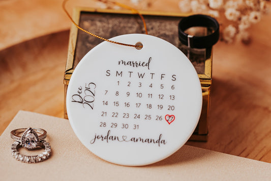 The Mirabilia Boutique custom ceramic ornament for couples features a December 2025 calendar with "Married" on top, highlighting the 20th with a heart. It reads "jordan & amanda," with wedding rings and a box in the backdrop, creating a special personalized keepsake.