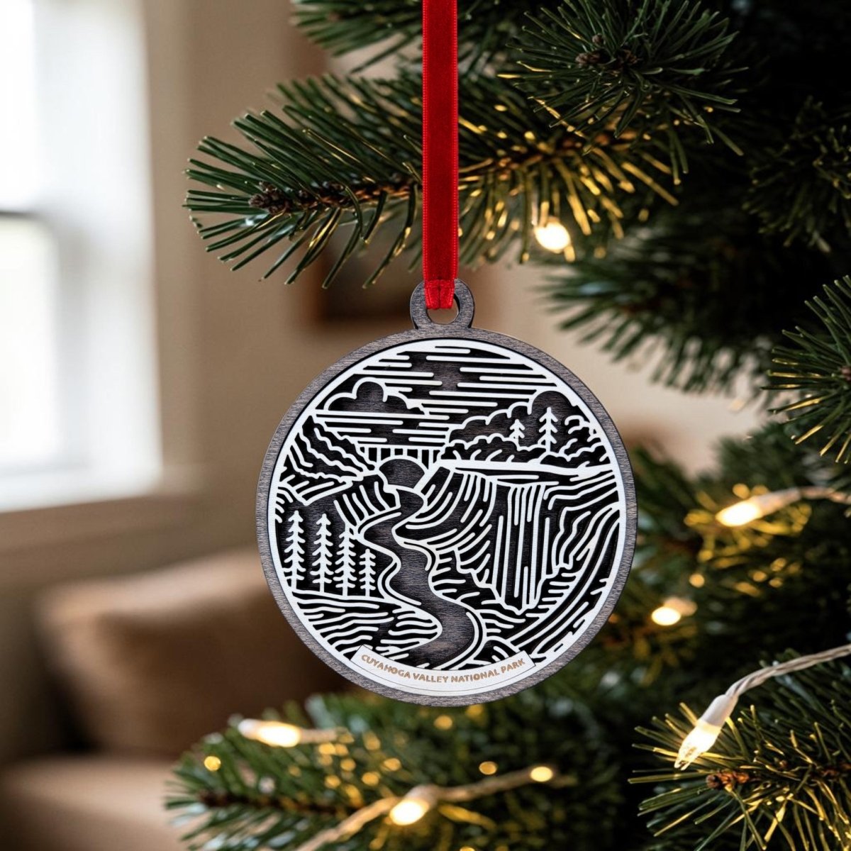 A Cuyahoga Valley National Park Christmas Ornament by Mirabilia Boutique hangs from a red ribbon on the tree. This oval wooden ornament features a black and white landscape scene with a river, trees, and bridge, and "Cuyahoga Valley National Park" inscribed at the bottom—capturing the serene beauty of this cherished destination.