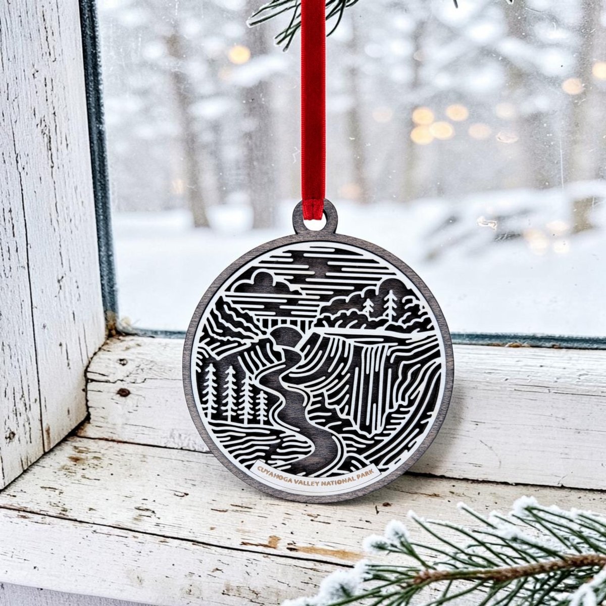 A Mirabilia Boutique "Cuyahoga Valley National Park Christmas Ornament" featuring a stylized river, trees, and waterfall—evoking Brandywine Falls—hangs from a red ribbon on a snowy window. The ornament displays "Cuyahoga Valley National Park" at the bottom, with pine needles visible in the foreground.
