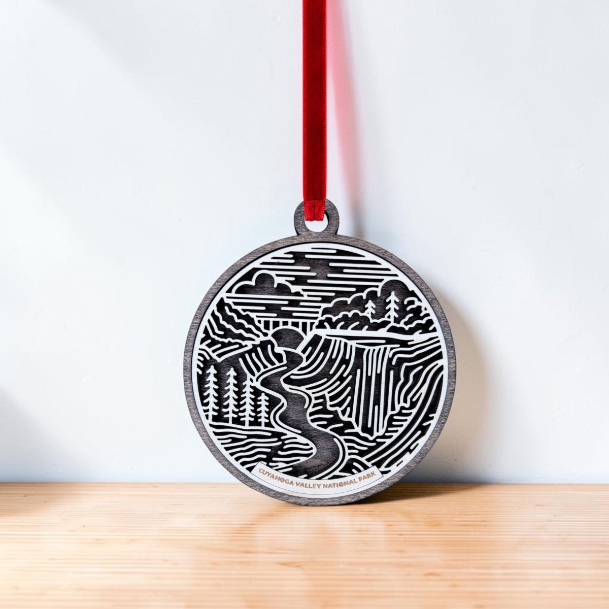 The Cuyahoga Valley National Park Christmas Ornament by Mirabilia Boutique—a round wooden decoration showcasing a nature scene with trees, a river, and a waterfall inspired by Brandywine Falls—hangs from a red ribbon against a light background, resting on a wooden surface.