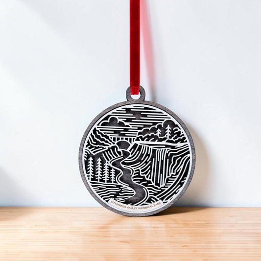 The Cuyahoga Valley National Park Christmas Ornament by Mirabilia Boutique—a round wooden decoration showcasing a nature scene with trees, a river, and a waterfall inspired by Brandywine Falls—hangs from a red ribbon against a light background, resting on a wooden surface.