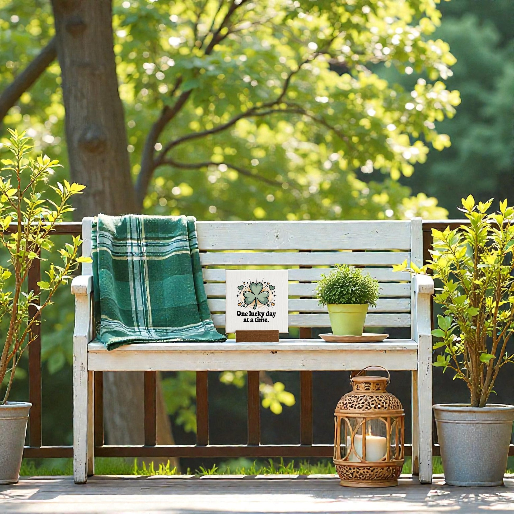 A white wooden bench on a sunny deck holds a green plaid blanket, potted plant, lantern, and the Mirabilia Boutique "One Lucky Day at a Time" Mini Sign (4x4), adding charming good fortune to the lush setting.