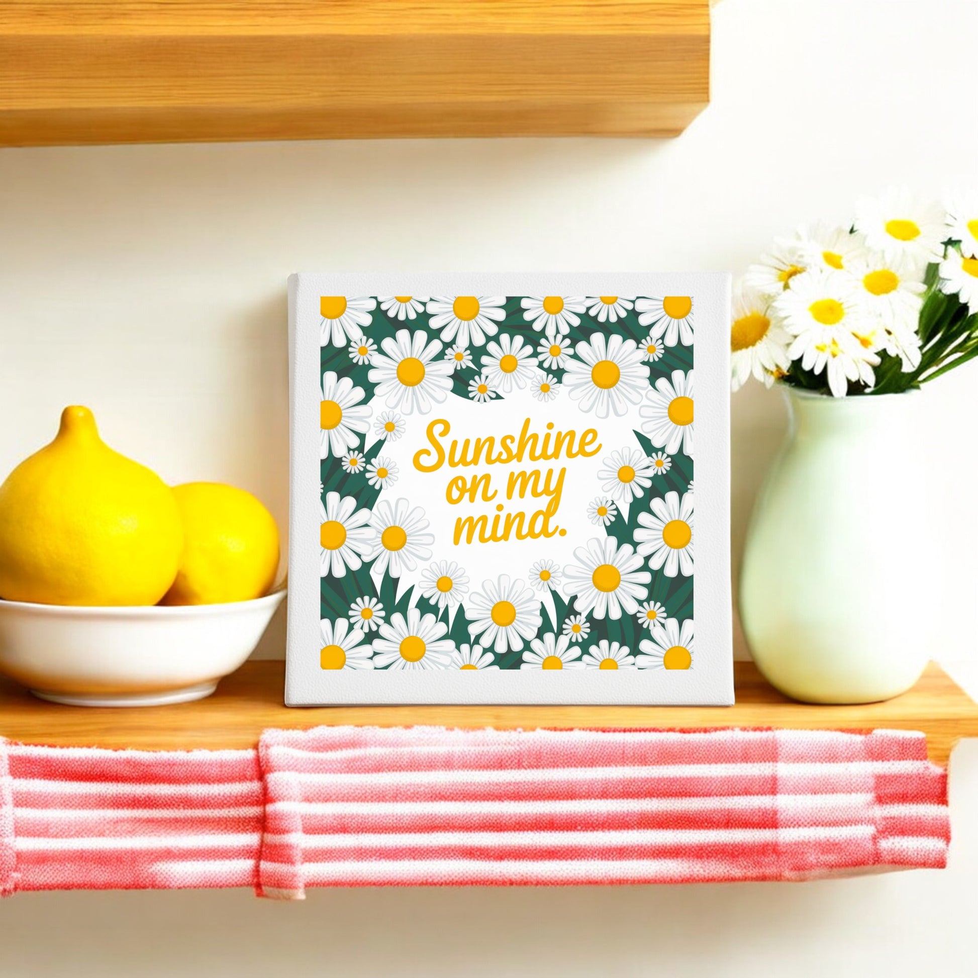 The Sunshine on My Mind Canvas – 8x8 Daisy Wall Art by Mirabilia Boutique sits beside lemons, daisies, and a striped cloth—perfect for brightening your home with cheerful summer decor.