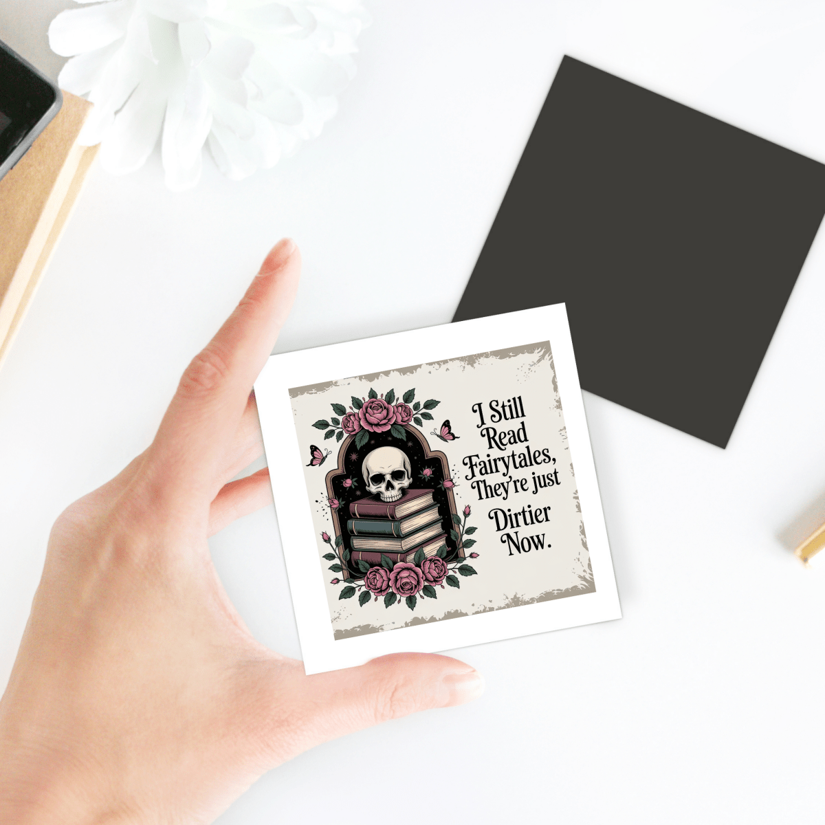 A hand holds the "I Still Read Fairytales Gothic Book Magnet" by Mirabilia Boutique, featuring a skull, roses, and books—an ideal literary gift. The magnet displays the phrase, "I Still Read Fairytales. They're just Dirtier Now." A black square and white flower add fairytale-inspired decor to the background on a white surface.