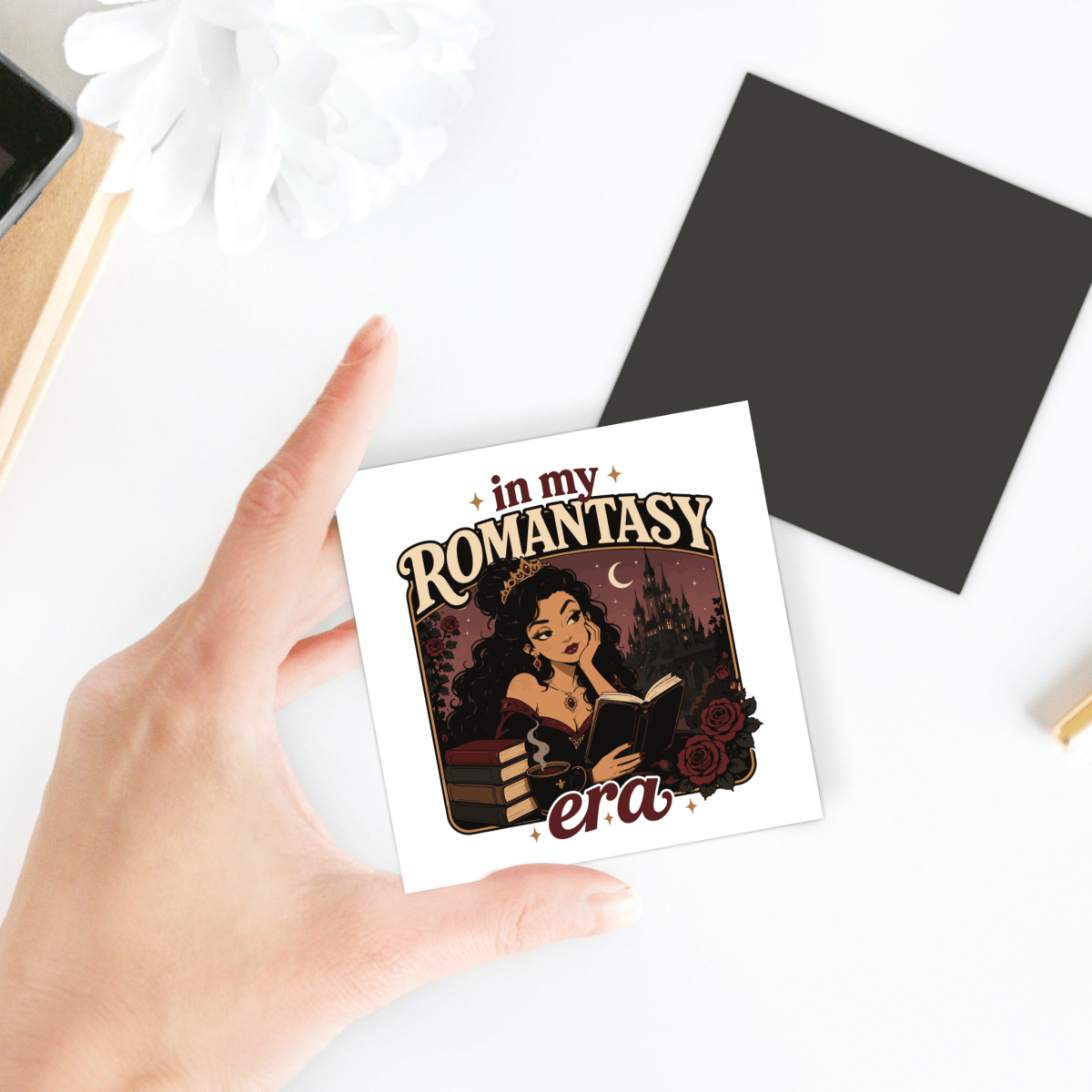 A hand holds the "In My Romantasy Era" magnet from Mirabilia Boutique—a perfect bookish accessory—featuring a smiling woman reading a book, with the text "in my ROMANTASY era" and roses on a dark, magical background. Ideal as a fantasy reader gift for those who adore mystical tales and enchanting aesthetics.