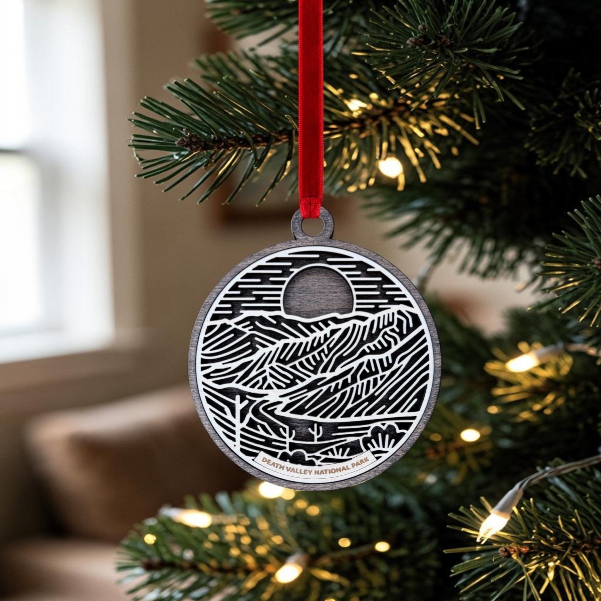 A Death Valley National Park Christmas Ornament from Mirabilia Boutique hangs from a red ribbon on a lit Christmas tree. This handcrafted wooden ornament features intricate line art of the park’s landscape with "Death Valley National Park" text, set against a blurred room in the background.