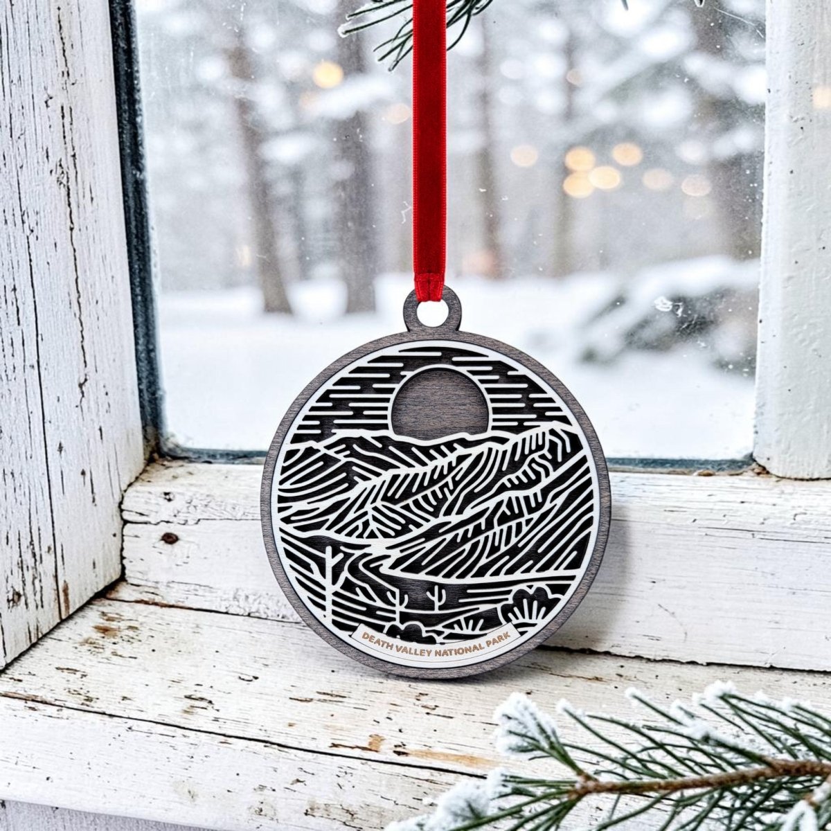 A handcrafted Death Valley National Park Christmas Ornament by Mirabilia Boutique hangs on a snowy window, featuring intricate wooden line art with a mountain design and "Death Valley National Park" engraved in silver. Suspended by a red ribbon, the ornament stands out against the view of pine branches and snow-covered trees outside, offering a unique tribute to this iconic desert landscape.