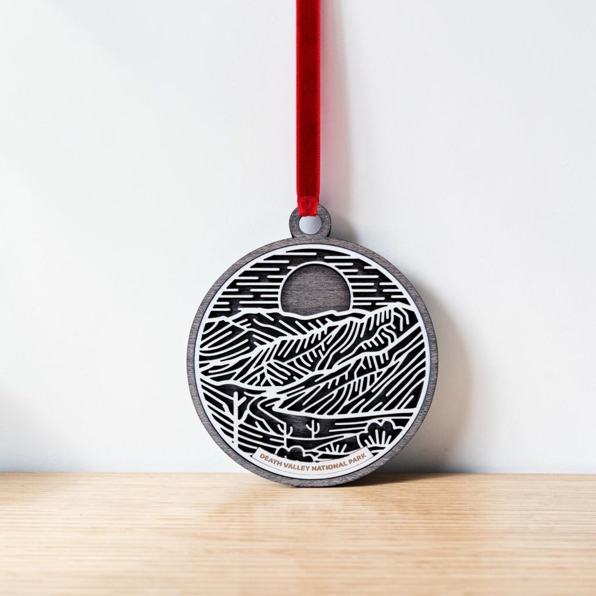 Celebrate the beauty of Death Valley with the Death Valley National Park Christmas Ornament by Mirabilia Boutique. This round metallic ornament features an engraved landscape of mountains, sun, and desert plants, accented by a vibrant red ribbon. "Death Valley National Park" is inscribed at the bottom, making it an ideal keepsake for desert landscape lovers and a perfect addition to your holiday décor. The ornament is pictured resting against a white wall.