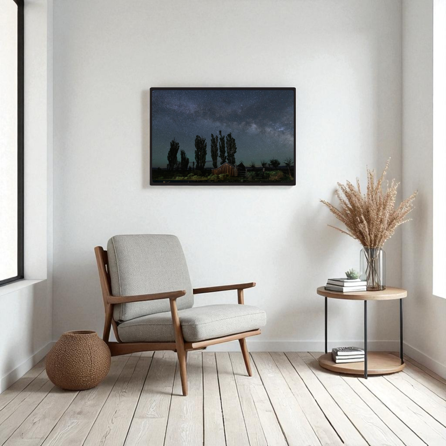 A modern, minimalist living room with a gray armchair, round side table adorned with dried grass, woven basket, and the DeChambeau Ranch Milky Way Print by Mirabilia Boutique—an archival-quality framed fine art print of the Eastern Sierra night sky—hanging on the wall above the chair.