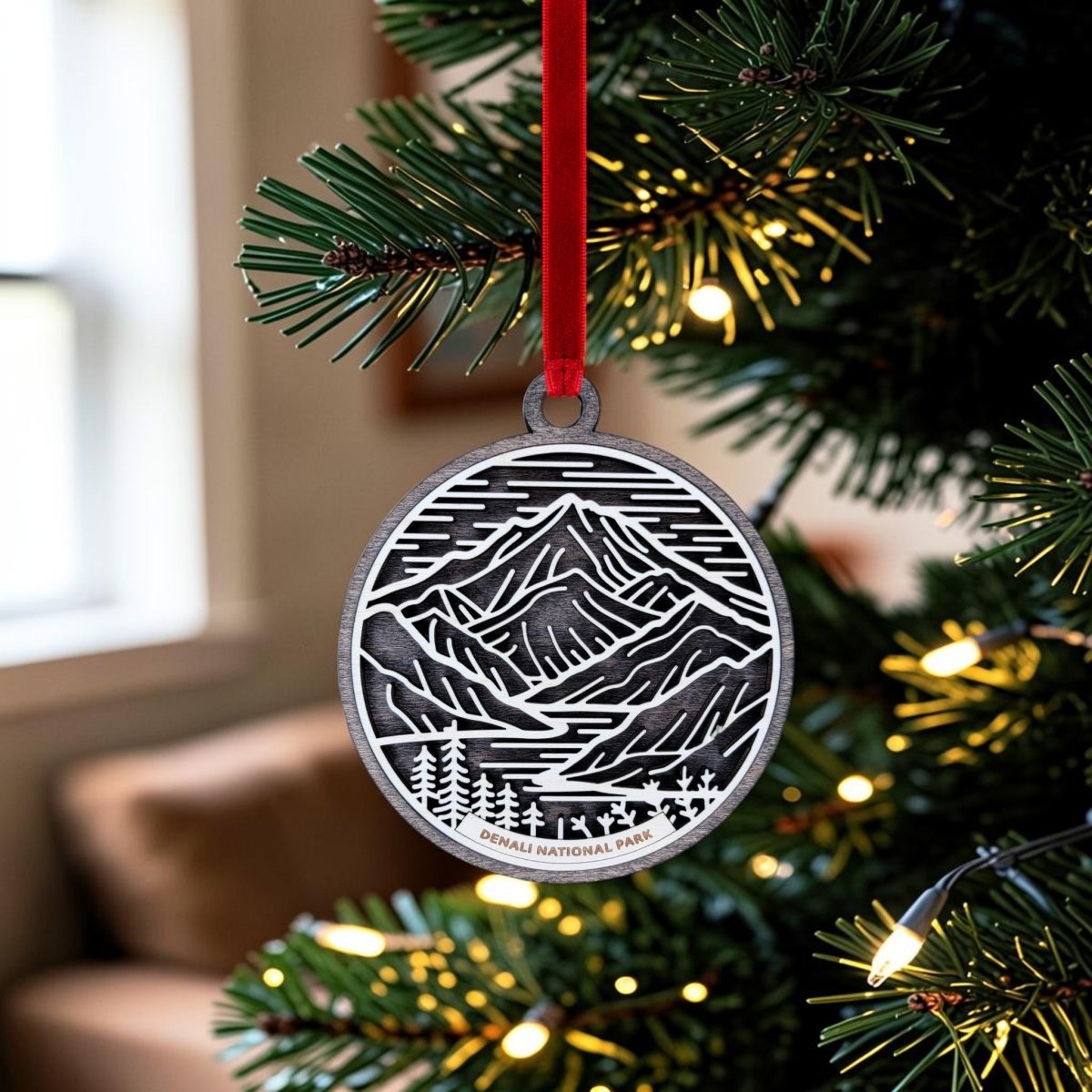 The Denali National Park Christmas Ornament by Mirabilia Boutique hangs from a vibrant red ribbon on a festive tree, its handcrafted wooden design showcasing a stunning Denali mountain scene amid twinkling lights and green branches—making it the perfect Alaska keepsake for the holiday season.