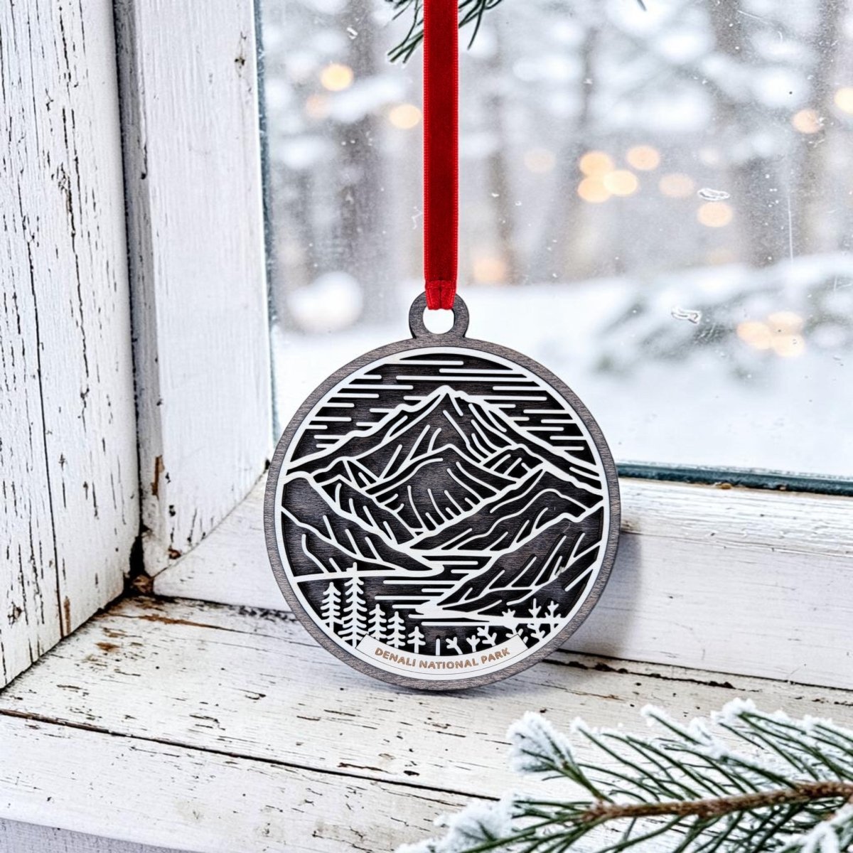 The Denali National Park Christmas Ornament by Mirabilia Boutique features a silver oval design with engraved mountains, trees, and "Denali National Park." Hanging from a red ribbon on a rustic white window frame, this Alaska keepsake is set against a snowy outdoor scene with a pine branch in the background—making it a charming addition to your holiday decor.