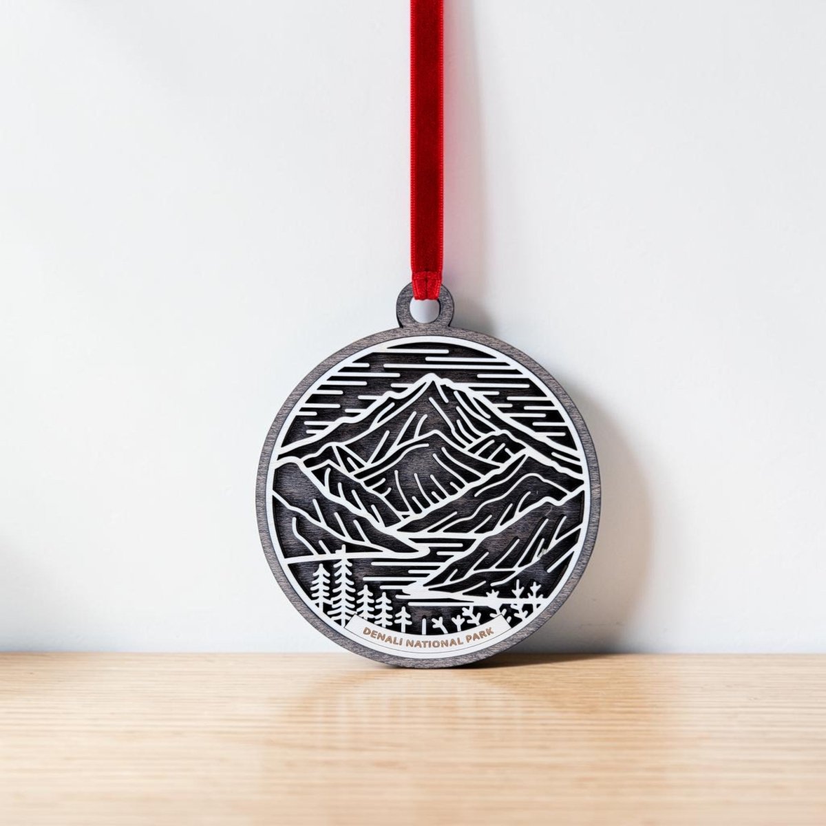 A handcrafted Mirabilia Boutique Denali National Park Christmas Ornament, featuring a mountain and pine tree design, hangs from a red ribbon against a white wall. "Denali National Park" is displayed at the bottom of this round wooden ornament, which rests on a light wooden surface—making it an ideal Alaska keepsake.