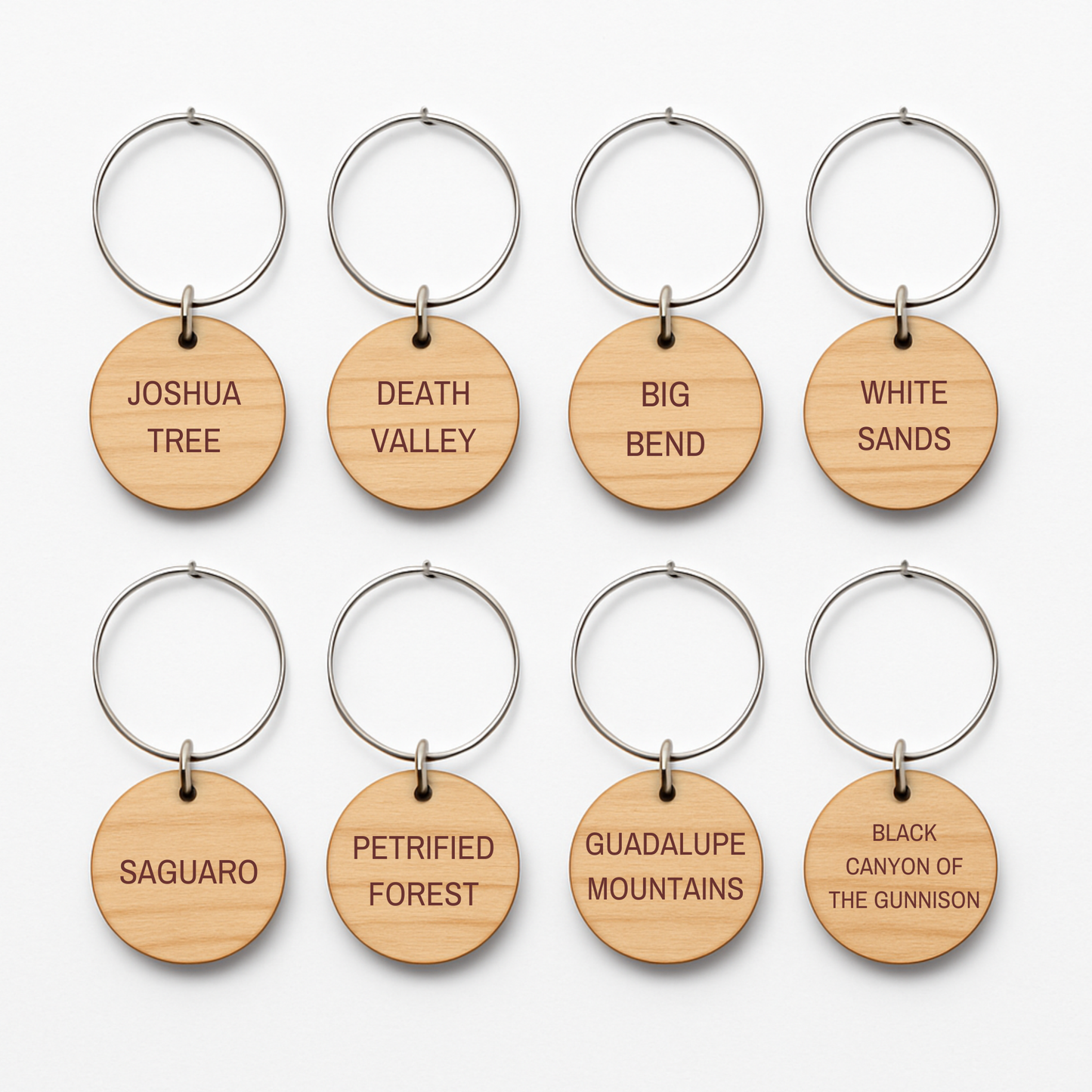 Eight round wooden wine charms from Mirabilia Boutique’s Desert Dreaming Wine Charms collection are arranged in two rows, each engraved with a different U.S. national park name like Joshua Tree, Death Valley, and White Sands.
