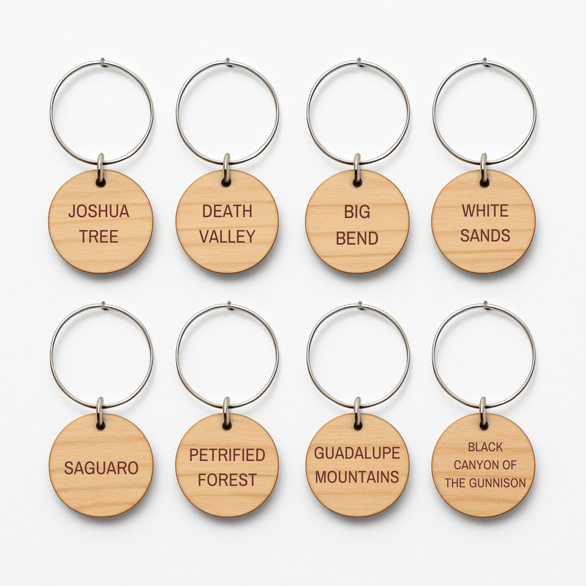 Eight round wooden wine charms from Mirabilia Boutique’s Desert Dreaming Wine Charms collection are arranged in two rows, each engraved with a different U.S. national park name like Joshua Tree, Death Valley, and White Sands.