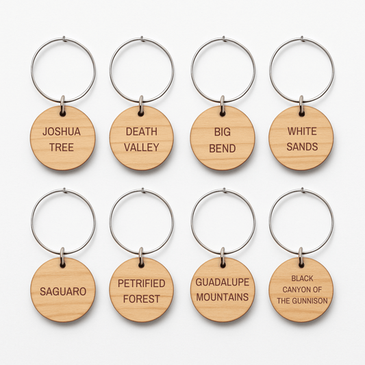 Eight round wooden wine charms from Mirabilia Boutique’s Desert Dreaming Wine Charms collection are arranged in two rows, each engraved with a different U.S. national park name like Joshua Tree, Death Valley, and White Sands.